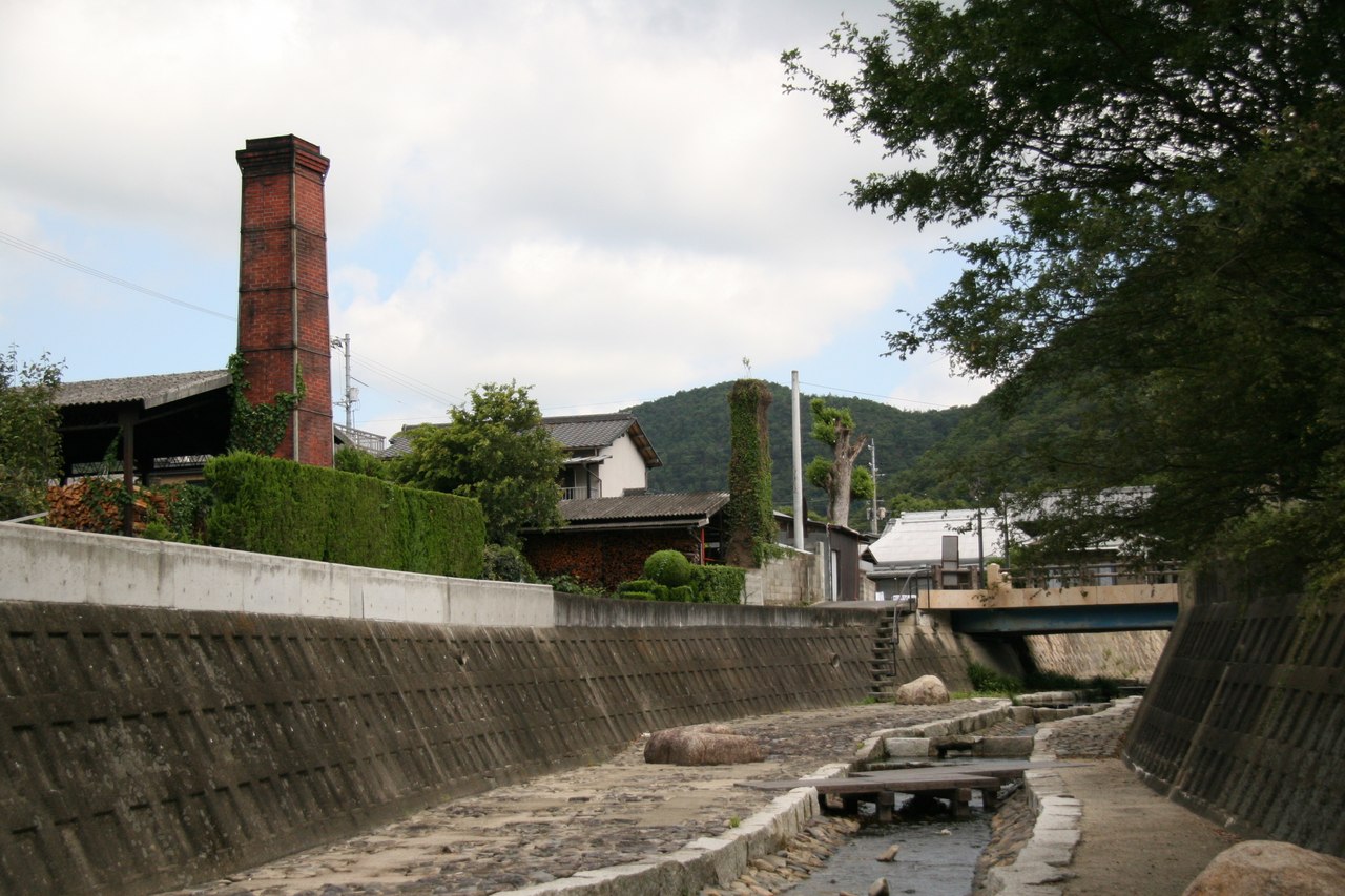 A tall brick kiln chimney rising above the workshops of Imbe, with stacked firewood below