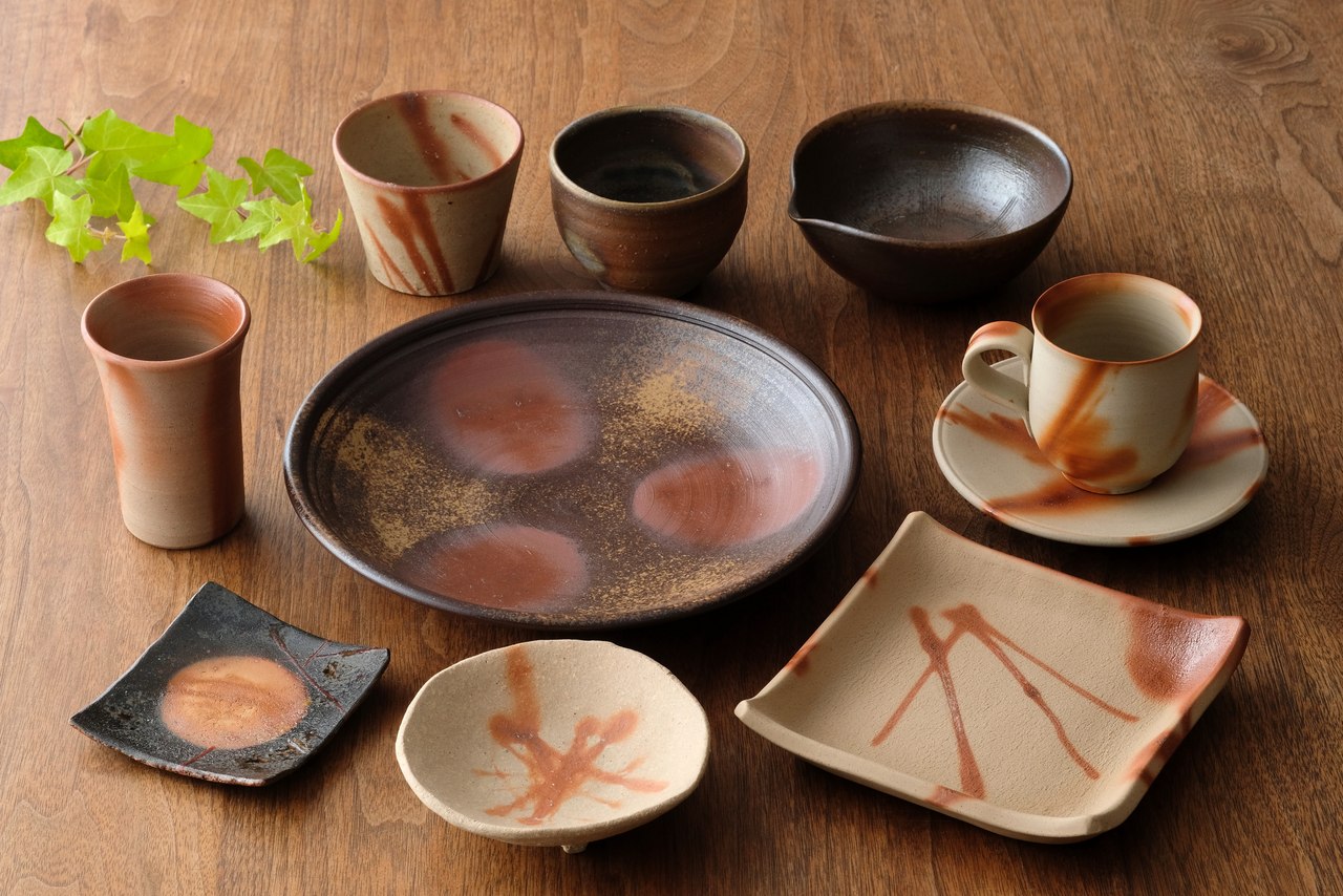 An assortment of Bizen ware on a wooden table — plates, small dishes, cups, a mug and saucer, and a pouring bowl