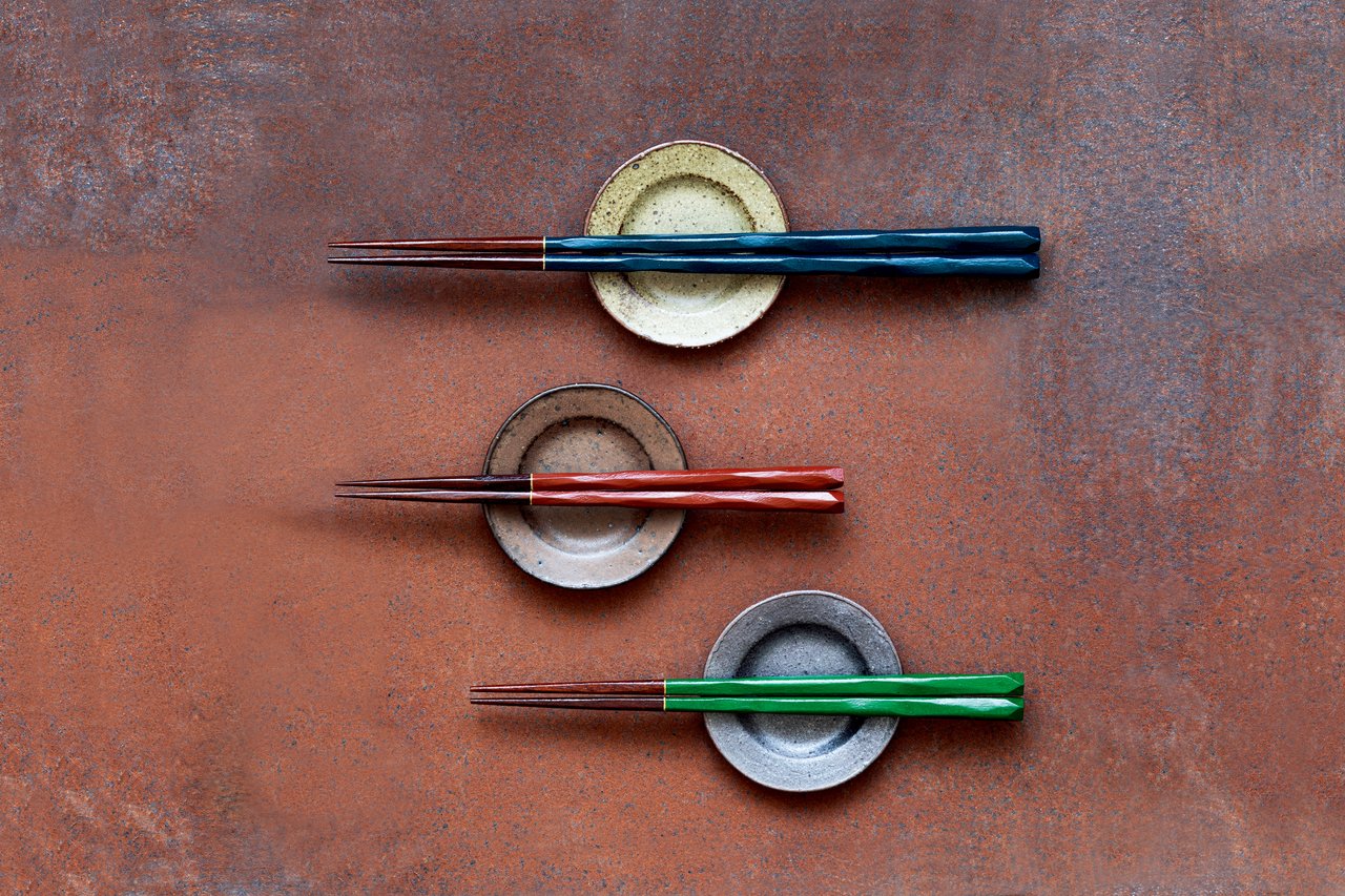 Three lacquered chopstick pairs resting on small ceramic hashioki rests against a rust-colored background