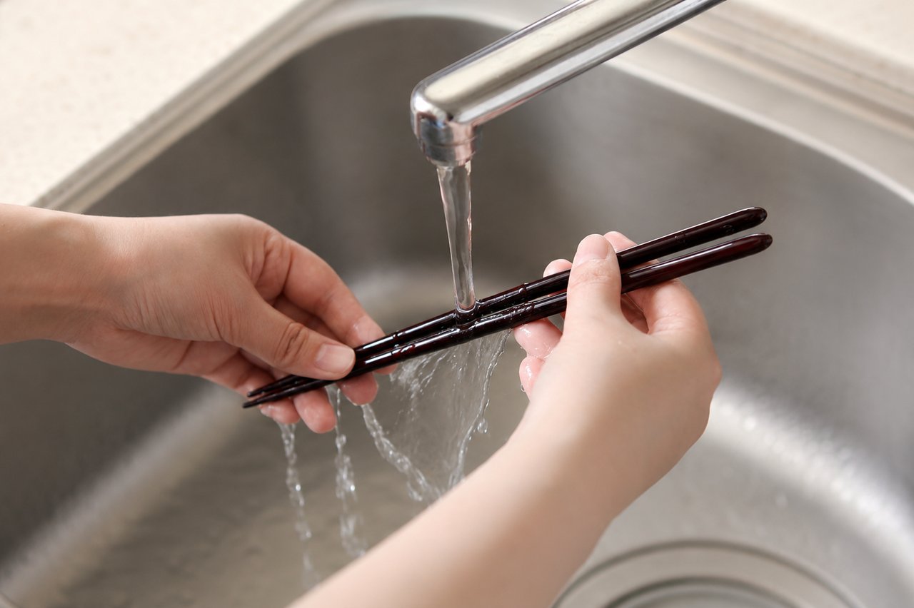 Two hands rinsing a pair of dark lacquered chopsticks under gently running water at a kitchen sink