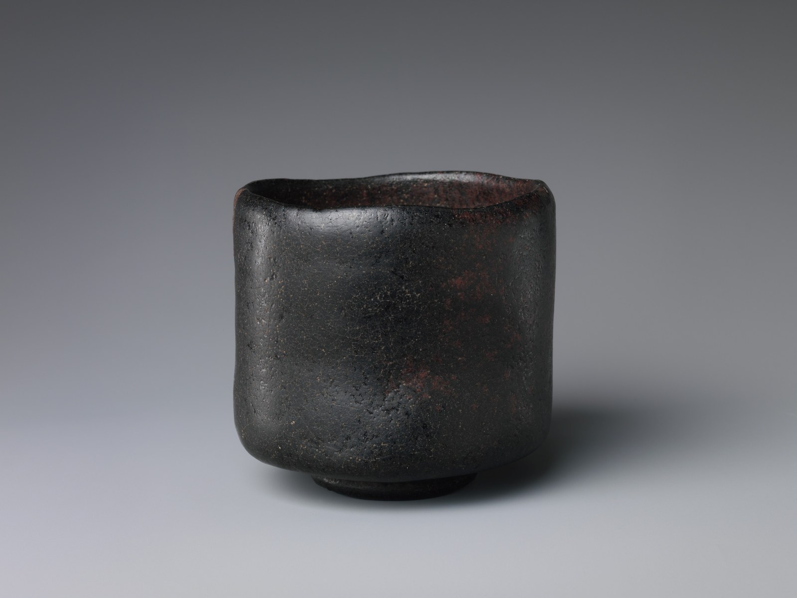 A tall black raku tea bowl with a softly uneven rim, against a gray background