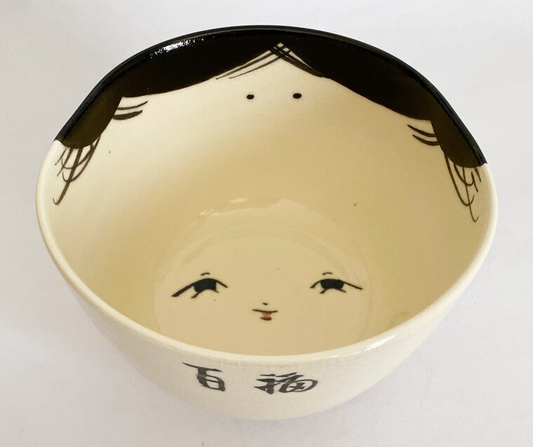 A cream-colored tea bowl painted inside with the smiling face of Okame, her black hair running along the rim
