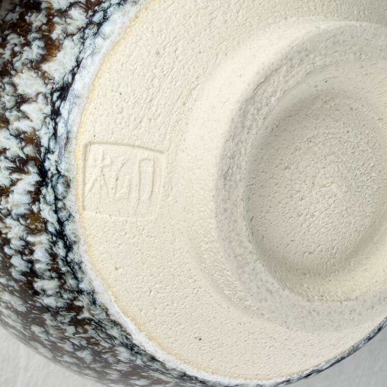 The underside of a tea bowl: an unglazed cream-colored foot ring with a small stamped seal, framed by a speckled glazed wall