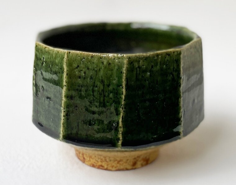 A tea bowl in deep green Oribe glaze, its wall cut into flat vertical facets, standing on an unglazed foot