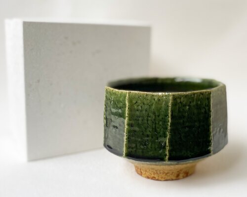 A faceted green Oribe tea bowl on an unglazed foot, beside a white box