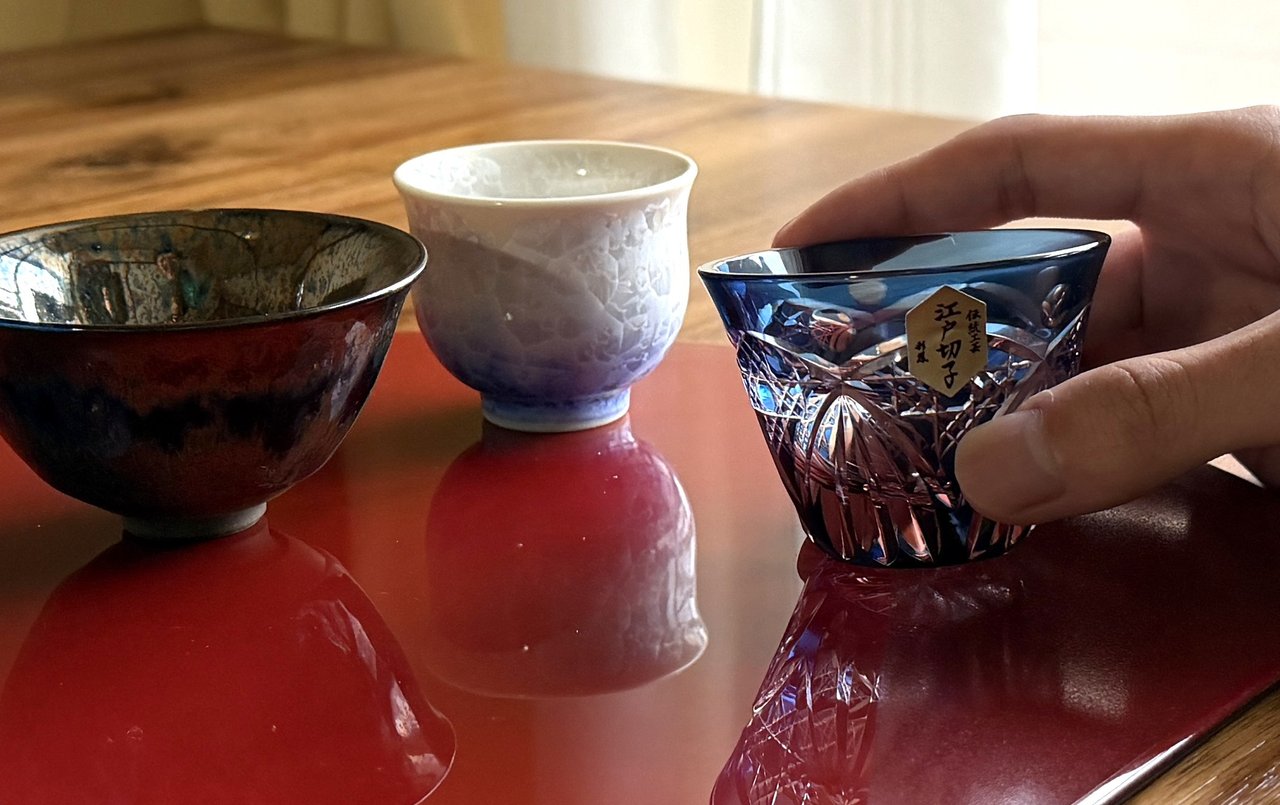 Three sake cups on a vermilion lacquer tray — a dark stoneware cup, a white porcelain cup, and a blue Edo Kiriko cut-glass guinomi — with a hand reaching toward the kiriko, the personal cup chosen for the evening