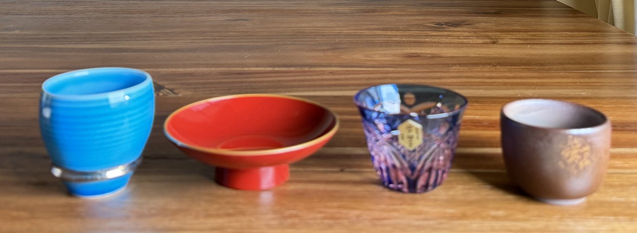 A vignette of multiple sake vessel materials side by side — porcelain, stoneware, tin, glass, and lacquer — each catching light differently