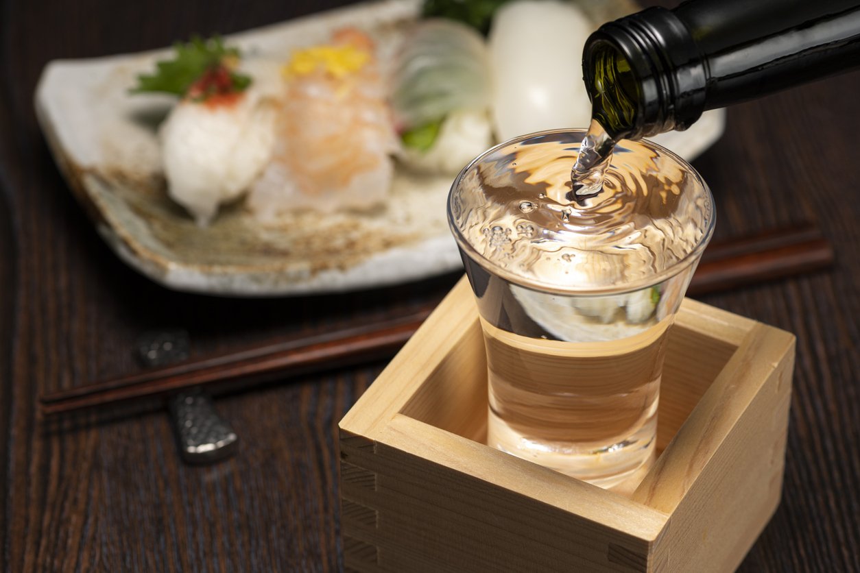 Sake overflowing from a glass into the cedar masu box beneath it, with sushi in the background