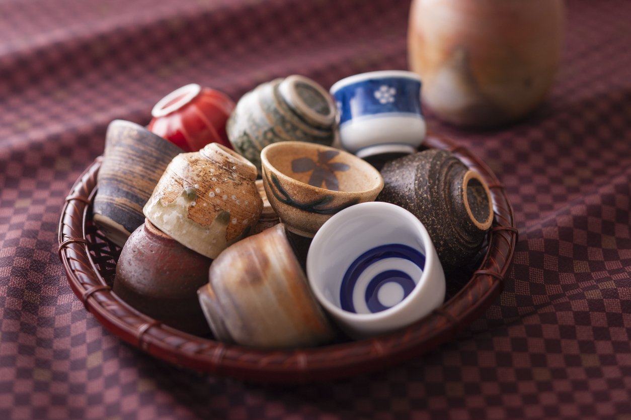 A woven basket filled with assorted Japanese sake cups in different glazes and patterns, including a janome tasting cup