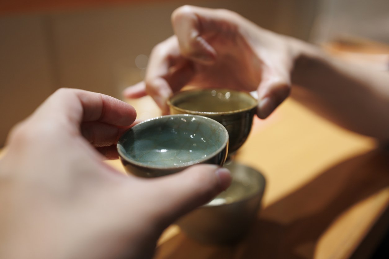 Two people gently touching small sake cups together over a warm izakaya table