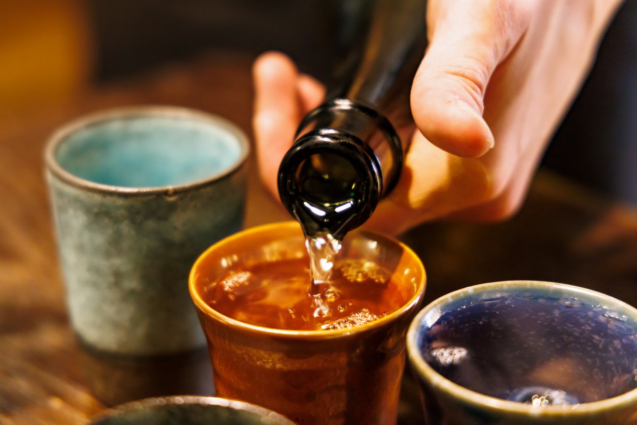 Sake being poured into a rustic amber cup beside cups in other glazes on a dark wooden table
