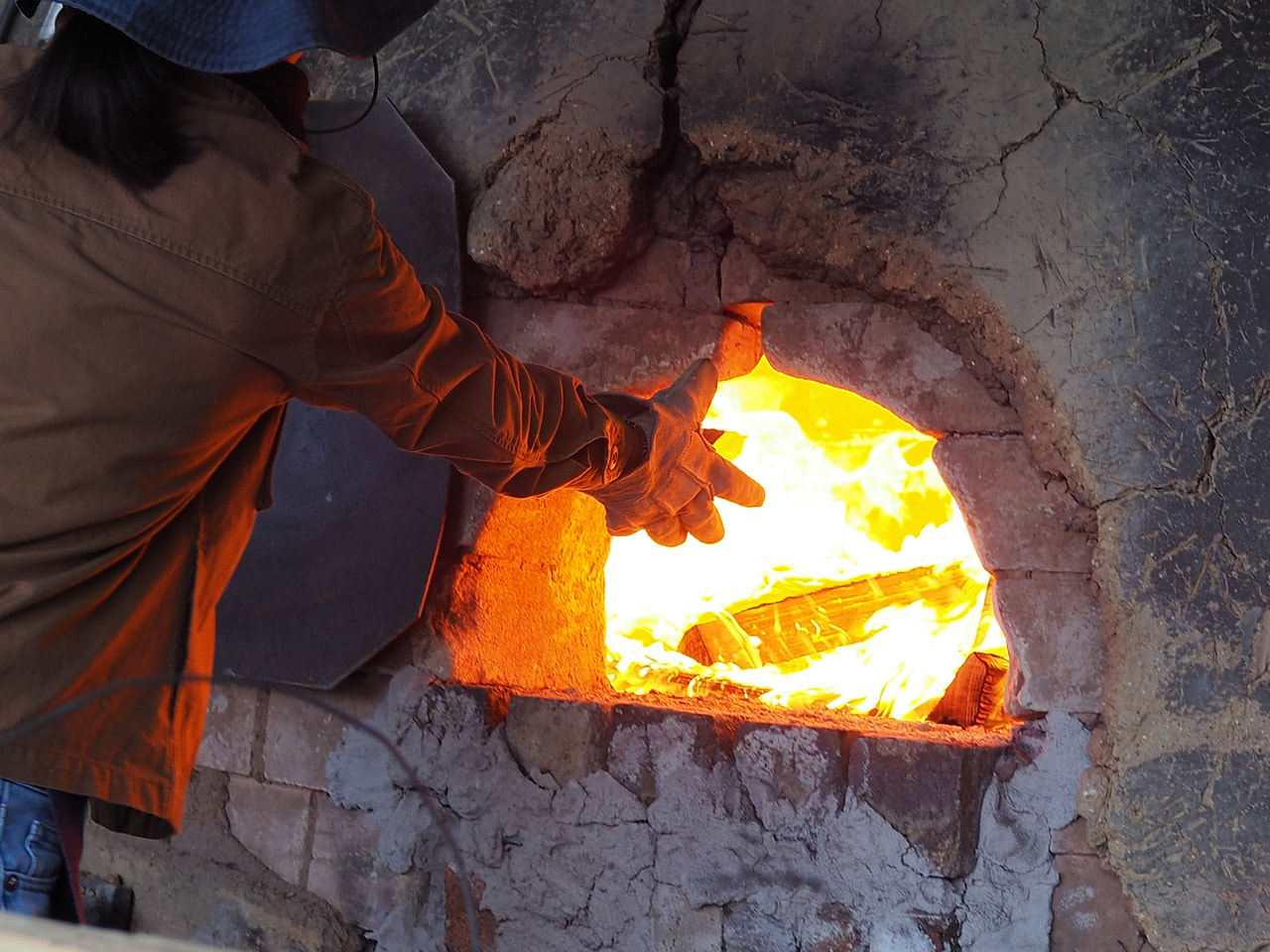 A potter tending the firebox of a wood-fired climbing kiln, flames visible through the stoking door