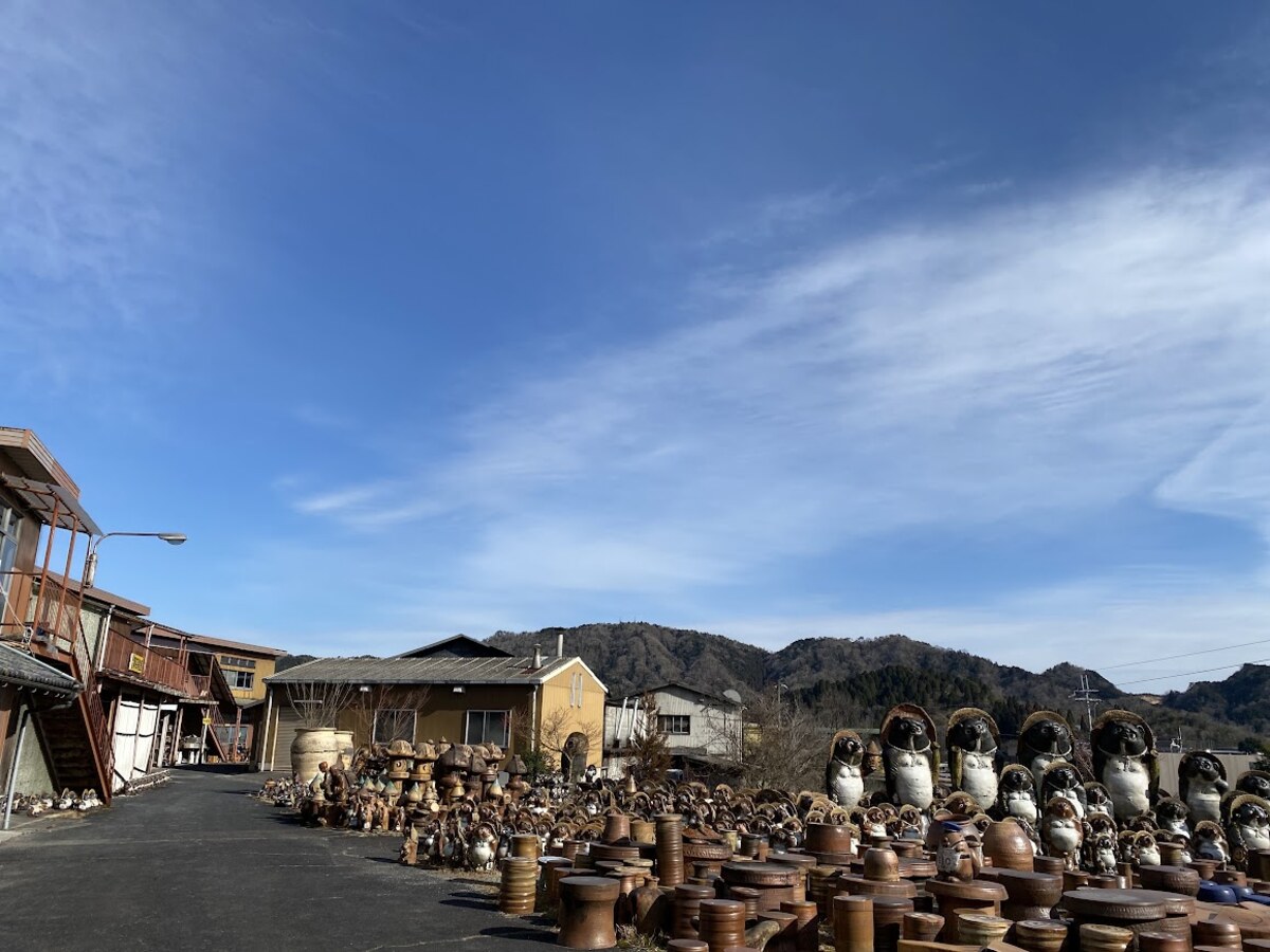Rows of tanuki statues for sale outside a shop along a Shigaraki street, with mountains in the background