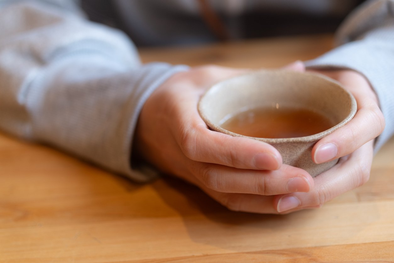 Two hands cradling a handleless Japanese teacup of hojicha on a wooden table