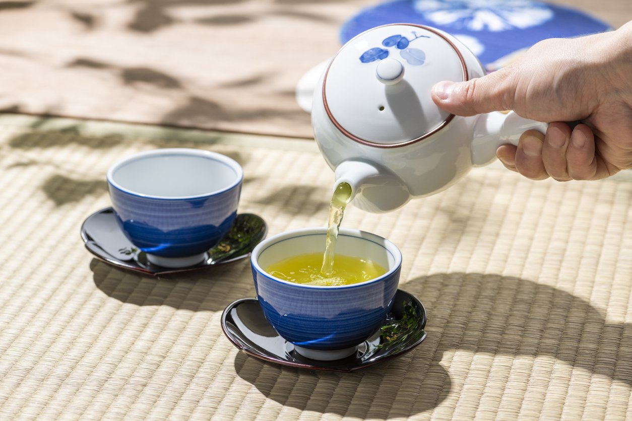 A Japanese teapot pouring bright green sencha into blue porcelain cups on a tatami mat