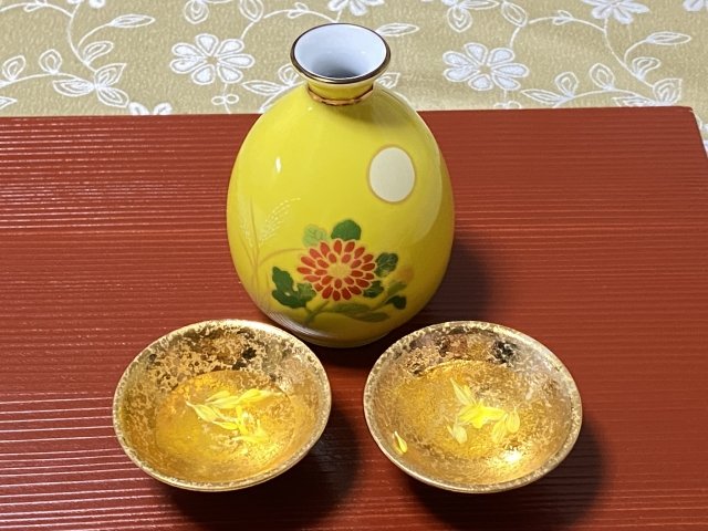 A yellow sake bottle painted with a chrysanthemum beside two gold-lined sake cups holding chrysanthemum petals