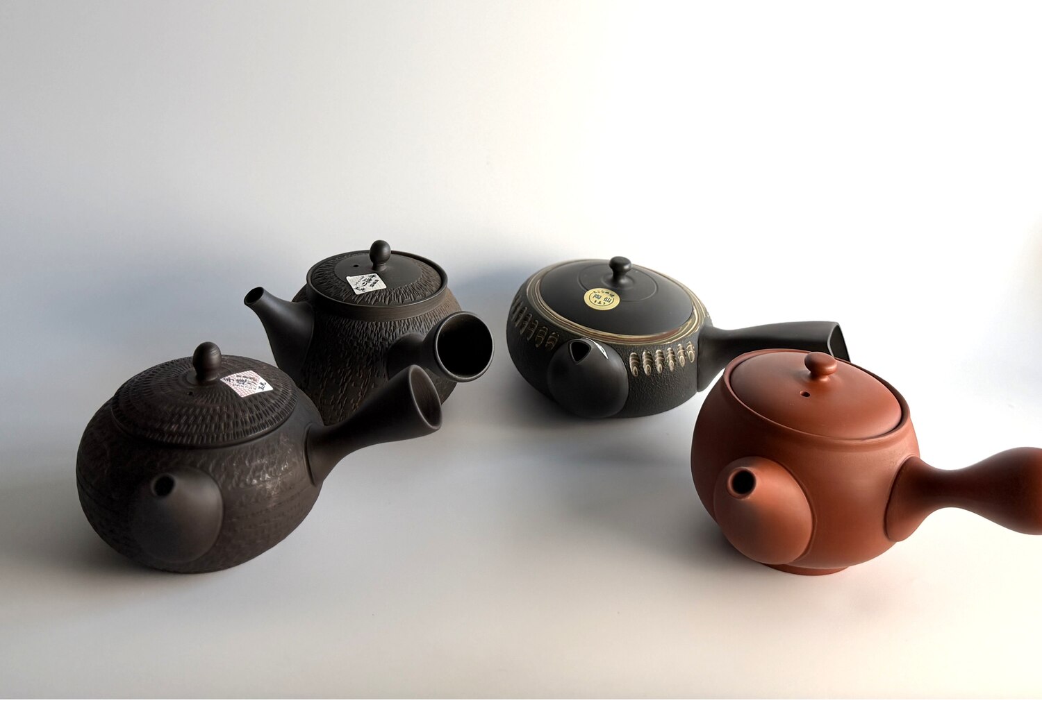 Four Tokoname teapots side by side, three dark unglazed clay and one vermilion shudei, showing a range of surface finishes