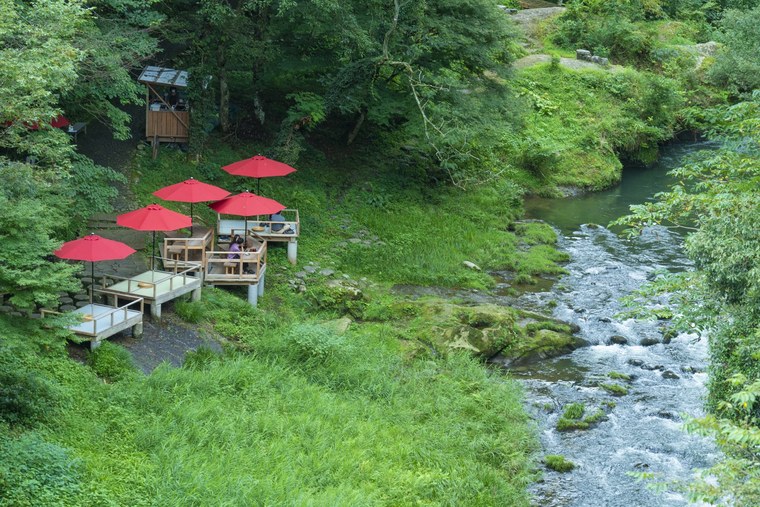A wooded gorge at Yamanaka Onsen with a clear stream running through it and riverside platforms shaded by red parasols