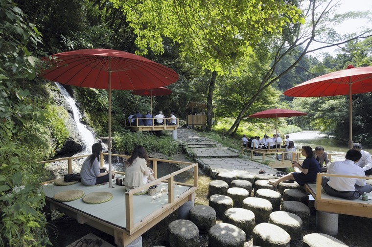 Wooden riverside platforms under red parasols beside a shallow stream in a wooded gorge, with visitors seated on them in summer
