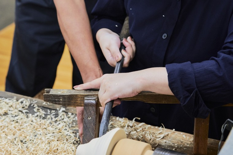 Two pairs of hands guiding a long chisel against a pale wooden blank turning on a lathe, with shavings gathered beside it
