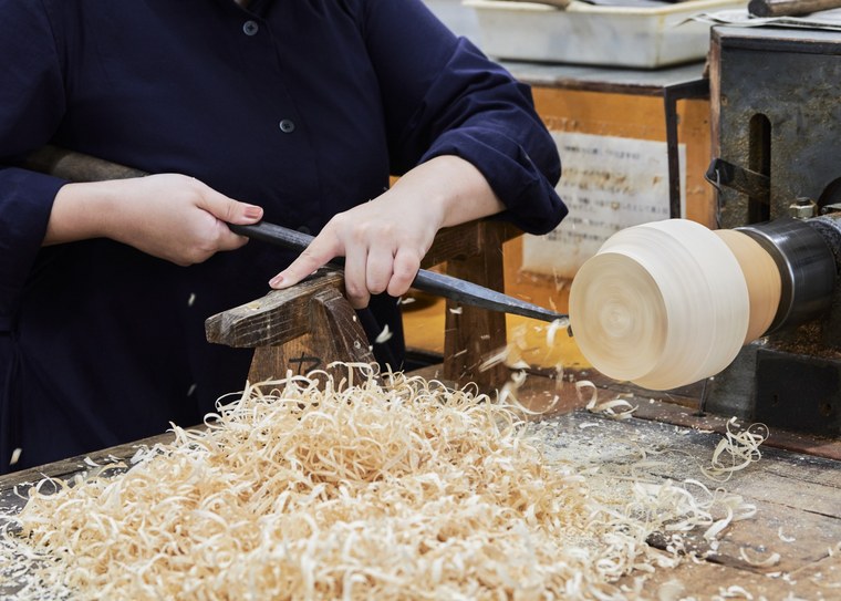 A craftsperson's hands holding a long chisel against a pale wooden blank turning on a lathe, with a deep pile of shavings below