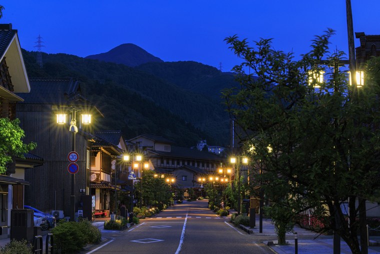 The main street of Yamanaka Onsen at dusk, lanterns lit along the road and a dark mountain ridge closing the view