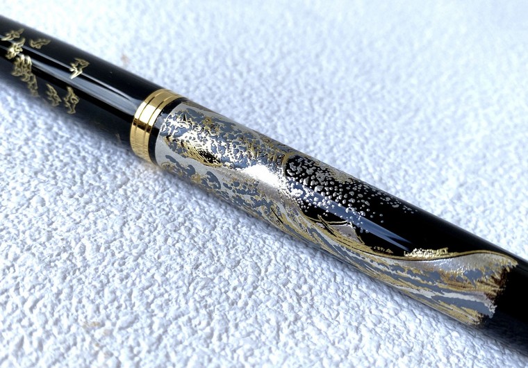 Close view of a black lacquered fountain pen barrel decorated with a breaking wave in gold and silver