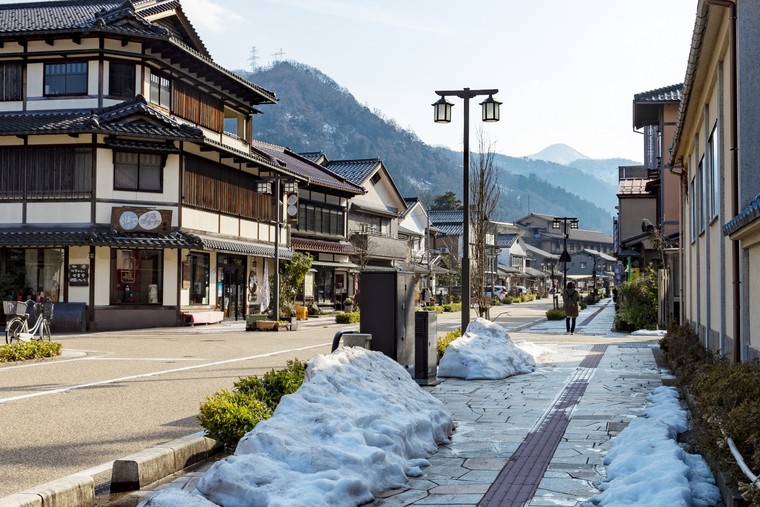 The main street of Yamanaka Onsen in late winter, snow banked along the pavement, wooden inns on either side and mountains beyond