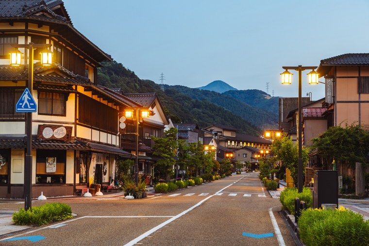 A street of low wooden inns and shops in Yamanaka Onsen at dusk, lit by lanterns, with mountains closing the far end