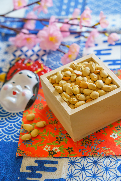 Roasted soybeans in a wooden masu box beside an oni mask and cherry blossoms&mdash;Setsubun in Japan.