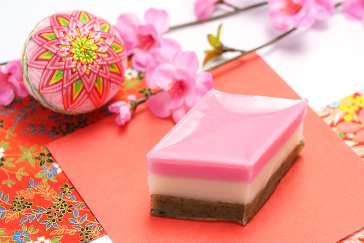 Hinamatsuri-style Japanese sweet with pink and white layers, arranged with cherry blossoms and colorful traditional fabric.
