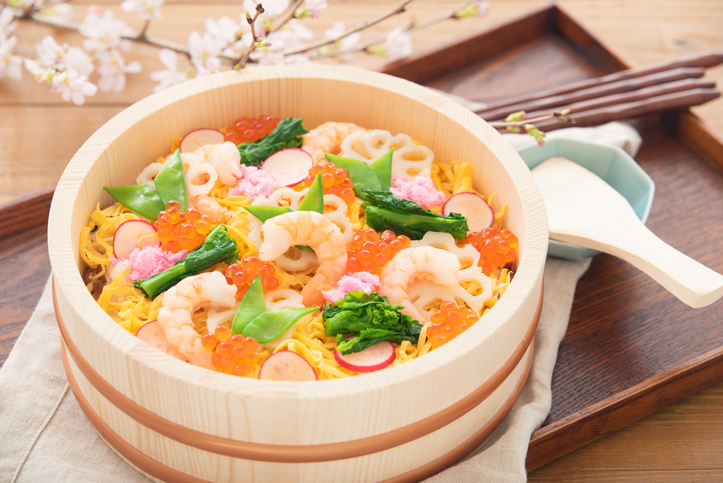 Chirashi sushi in a wooden bowl topped with shrimp and salmon roe, styled with spring cherry blossoms on a wooden table.