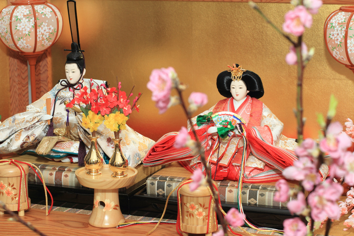 Traditional Japanese hina doll display with ornate kimono, ceremonial accessories, and pink peach blossoms in the foreground.